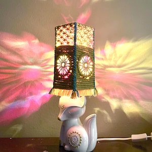 May include: A table lamp featuring a white ceramic fox base with gold accents and a square lampshade made of olive green macrame cord. The shade has a lattice pattern, circular floral motifs, and a fringe trim, casting colorful light patterns.