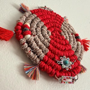 May include: A circular macrame wall hanging featuring alternating spiral coils of red and tan cord. The piece is decorated with small wooden beads, a silver-toned flower charm with a turquoise center, a rectangular bead frame, and multi-colored tassels.