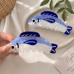 以下が含まれることがあります： 青と白のマーブル模様のアクリル製魚型ヘアクリップ2点。各クリップには、濃い青色の背びれ、濃い青色の尾、黒い瞳の白い目が付いています。本体は上部が水色、下部が白色で構成されています。