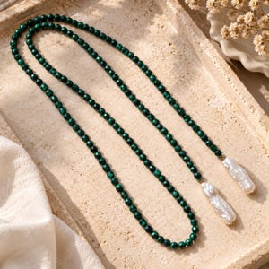 May include: A long necklace featuring a continuous strand of small, faceted dark green beads. The two ends of the necklace are finished with elongated, irregular white baroque pearls, each attached to the green bead strand with a small gold-toned metal cap.