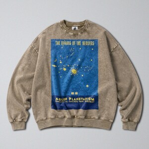 May include: A brown acid-wash crewneck sweatshirt featuring a large blue rectangular graphic on the chest. The graphic depicts a yellow sun, stars, and the text "THE DRAMA OF THE HEAVENS" above "ADLER PLANETARIUM OPERATED BY CHICAGO PARK DISTRICT."