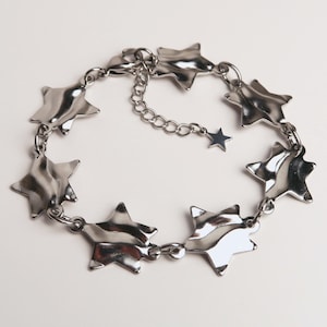 May include: A silver-toned metal bracelet featuring a series of connected star-shaped charms with a smooth, reflective surface. The bracelet includes a delicate chain extension that terminates in a small, singular star charm at the end.