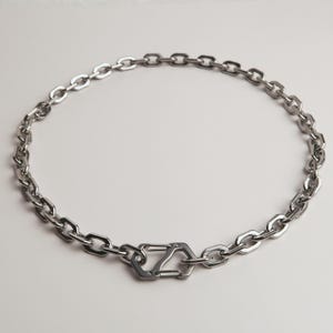 Puede incluir: Un collar de cadena plateada con un cierre geométrico único. El collar presenta una serie de eslabones de forma ovalada conectados a un cierre central con un diseño distintivo. La joya se muestra sobre un fondo blanco liso.