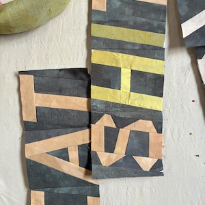 Könnte beinhalten: Ein Quiltblock-Ensemble aus Stoffstreifen in Anthrazit, blassem Pfirsich und Limettengrün. Das geometrische Muster besteht aus rechteckigen und dreieckigen Stoffstücken, die zusammengenäht wurden, um Buchstaben und Formen zu bilden.