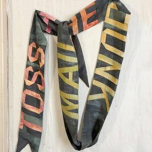 Op de afbeelding: Een donkergrijze sjaal met de woorden "LOSE" en "FUNK" in oranje, geel en roze. De sjaal is geknoopt en hangt tegen een witte achtergrond. De tekst is in een vette, blokkerige letter.