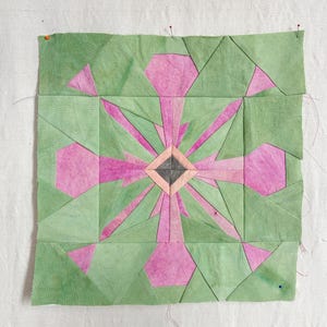 May include: A square quilt block featuring a geometric star pattern constructed from fabric pieces in shades of sage green, magenta, and soft pink. The center is a small dark gray diamond surrounded by light pink, radiating into a starburst design.
