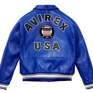 Puede incluir: Una chaqueta de cuero azul brillante con cuello cl&aacute;sico y mangas largas. La parte posterior muestra el texto AVIREX en letras negras, un parche circular con un dise&ntilde;o de bandera estadounidense y el texto USA en letras negras con bordes blancos.