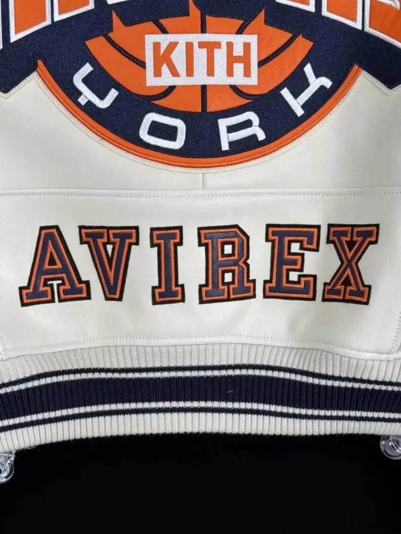 May include: A white jacket featuring a large circular orange and navy blue patch with the text KITH and YORK. Below the patch, the word AVIREX is embroidered in large orange letters with navy blue outlines. The jacket has a ribbed hem with navy stripes.