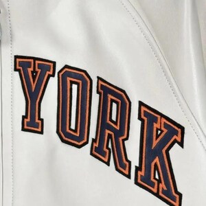 May include: A white jacket featuring the word YORK in large, block-style letters. The letters are dark blue with a thick orange outline and a thin black border, stitched onto the smooth white surface of the garment, which includes a collar.