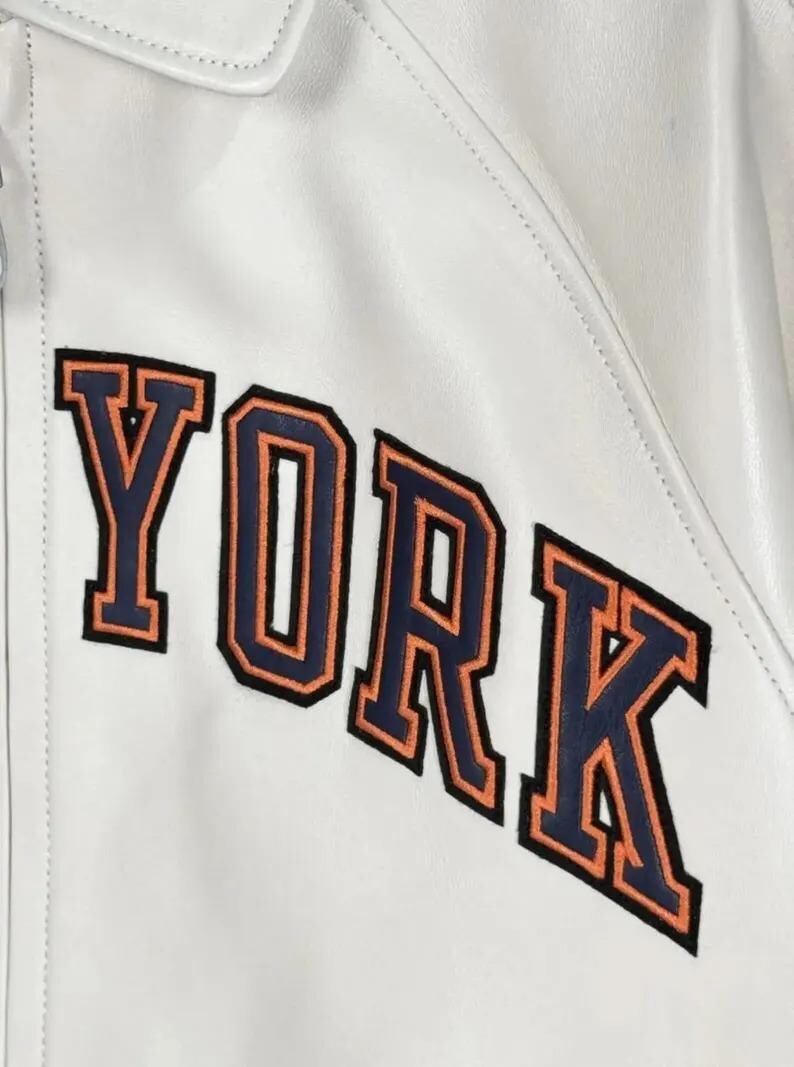 May include: A white jacket featuring the word YORK in large, block-style letters. The letters are dark blue with a thick orange outline and a thin black border, stitched onto the smooth white surface of the garment, which includes a collar.