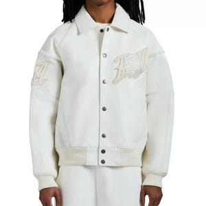 May include: A cream-colored varsity jacket featuring a pointed collar, snap-button front closure, and ribbed cuffs and hem. The jacket displays textured off-white patches on the chest and sleeves, with mesh-like fabric panels on the upper arms.