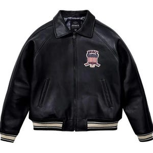 May include: A black leather jacket featuring a front zipper closure, a pointed collar, and ribbed cuffs and hem with white stripes. The chest displays an embroidered patch with the text AVIREX and a graphic of the American flag shield.