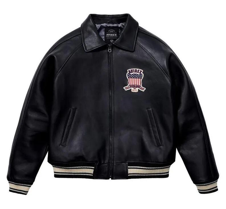 May include: A black leather jacket featuring a front zipper closure, a pointed collar, and ribbed cuffs and hem with white stripes. The chest displays an embroidered patch with the text AVIREX and a graphic of the American flag shield.