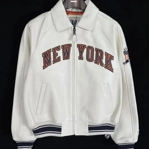May include: A white leather jacket featuring a full-length front zipper, a classic collar, and ribbed cuffs and hem with navy blue and white stripes. The chest displays the text NEW YORK in large, dark-outlined letters, with an Avirex logo on the sleeve.
