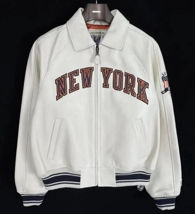 May include: A white leather jacket featuring a full-length front zipper, a classic collar, and ribbed cuffs and hem with navy blue and white stripes. The chest displays the text NEW YORK in large, dark-outlined letters, with an Avirex logo on the sleeve.
