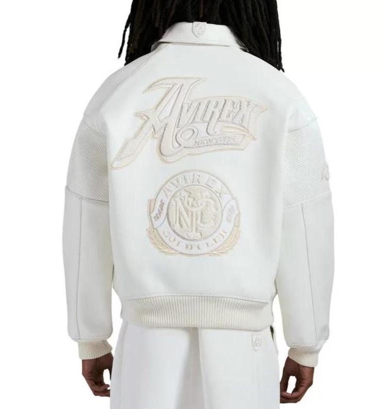 May include: A white varsity-style jacket featuring a large embroidered Avirex New York logo on the back. Below the text is a circular patch with the text Avirex Gold Club NYC. The jacket has ribbed cuffs and hem, with textured panels on the sleeves.