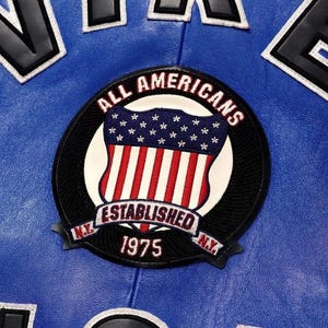 Puede incluir: Una chaqueta de cuero azul con un parche bordado circular. El parche muestra un escudo con estrellas y rayas, el texto ALL AMERICANS, ESTABLISHED 1975 y N.Y. en la parte inferior. Se ven letras negras grandes con borde blanco arriba.