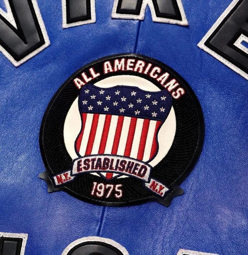 Puede incluir: Una chaqueta de cuero azul con un parche bordado circular. El parche muestra un escudo con estrellas y rayas, el texto ALL AMERICANS, ESTABLISHED 1975 y N.Y. en la parte inferior. Se ven letras negras grandes con borde blanco arriba.