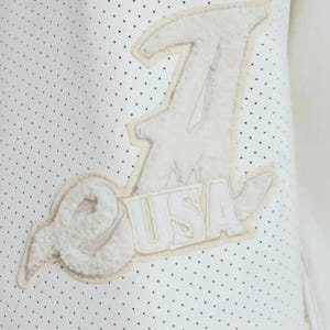 May include: A white perforated garment featuring a large, textured, off-white chenille patch. The patch displays a stylized letter A, a circular emblem, and the text USA in block letters, all stitched onto the surface with a light border.