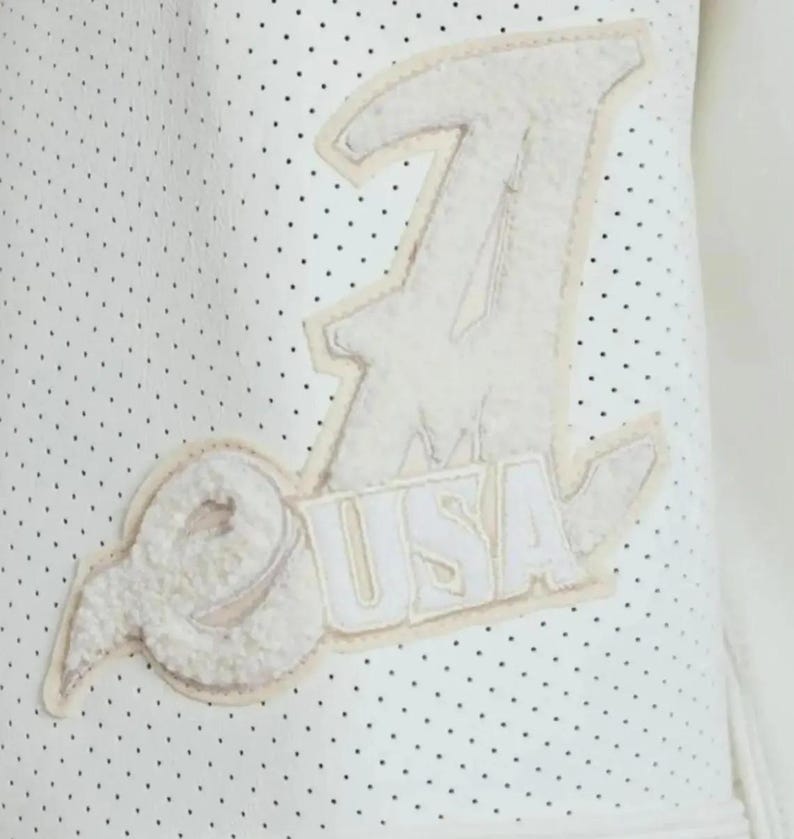 May include: A white perforated garment featuring a large, textured, off-white chenille patch. The patch displays a stylized letter A, a circular emblem, and the text USA in block letters, all stitched onto the surface with a light border.