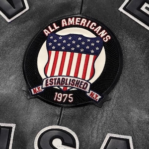 May include: A black leather jacket featuring a circular embroidered patch with the text ALL AMERICANS ESTABLISHED 1975 N.Y. N.Y. and a shield graphic with stars and stripes. Additional black leather lettering with white borders is visible.