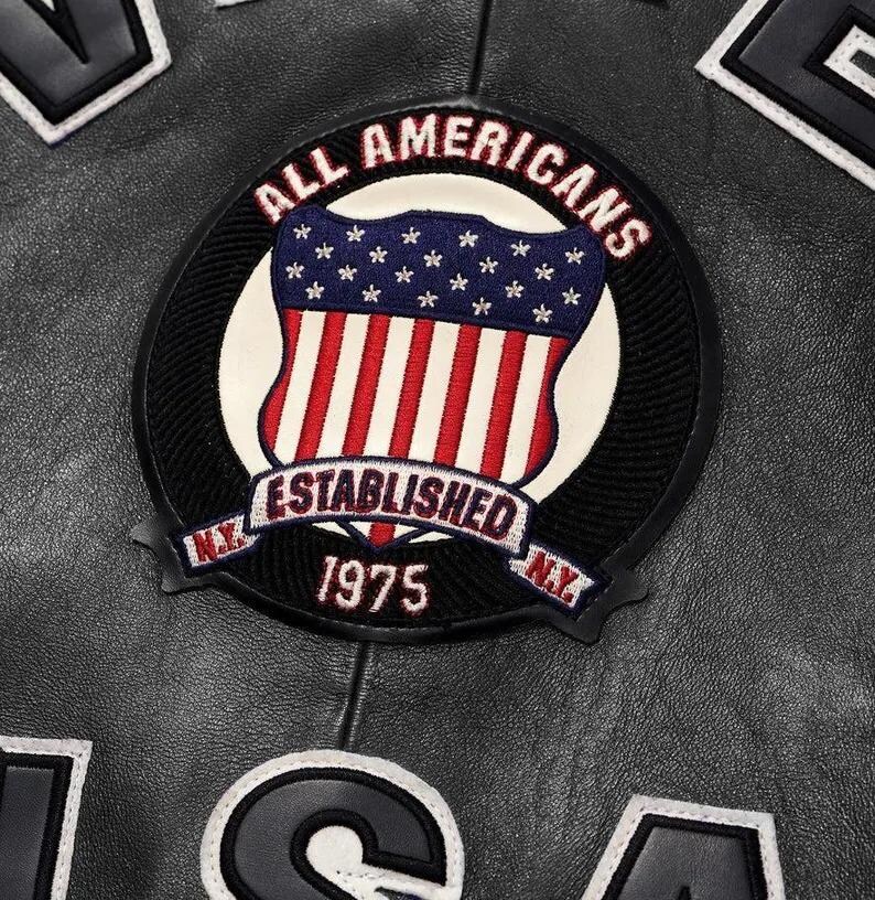 May include: A black leather jacket featuring a circular embroidered patch with the text ALL AMERICANS ESTABLISHED 1975 N.Y. N.Y. and a shield graphic with stars and stripes. Additional black leather lettering with white borders is visible.
