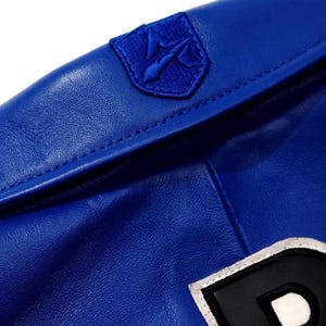 Puede incluir: Una chaqueta de cuero azul vibrante con un parche bordado en forma de escudo en el cuello. El parche muestra un s&iacute;mbolo de tridente estilizado en hilo azul a juego. Una parte de un parche de letra en blanco y negro es visible en la parte inferior.