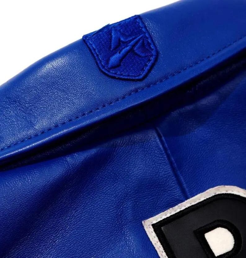 Puede incluir: Una chaqueta de cuero azul vibrante con un parche bordado en forma de escudo en el cuello. El parche muestra un s&iacute;mbolo de tridente estilizado en hilo azul a juego. Una parte de un parche de letra en blanco y negro es visible en la parte inferior.