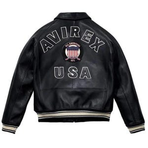 May include: A black leather varsity jacket featuring the text AVIREX in white arched lettering across the upper back. Below the text is a circular patch with an American flag design and the text ALL AMERICANS ESTABLISHED 1975. USA is printed below.