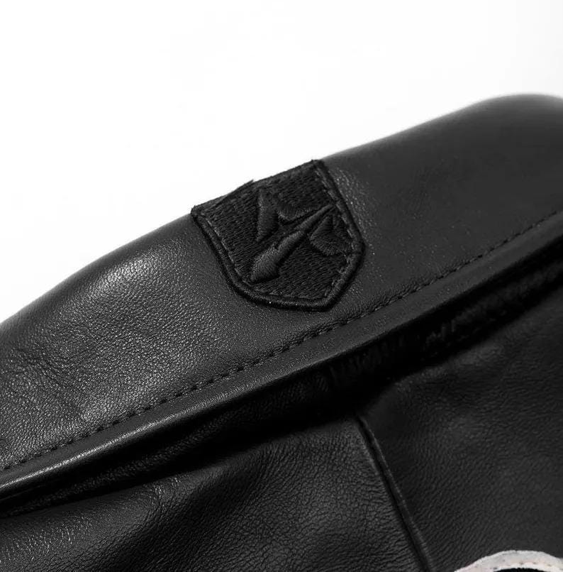 May include: A black leather accessory featuring a smooth texture and a prominent stitched seam along the top edge. An embroidered black patch with a stylized trident-like symbol is centered on the upper portion of the leather surface.
