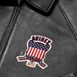 May include: A black leather jacket featuring a stitched patch on the chest. The patch displays a shield with red and white stripes and blue stars, the text AVIREX, and a banner reading N.Y. ESTABLISHED 1975 N.Y. in red and blue embroidery.