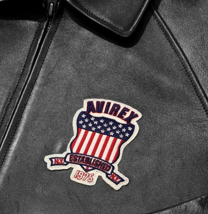 May include: A black leather jacket featuring a stitched patch on the chest. The patch displays a shield with red and white stripes and blue stars, the text AVIREX, and a banner reading N.Y. ESTABLISHED 1975 N.Y. in red and blue embroidery.