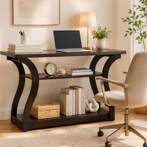 May include: A dark brown wooden desk featuring a curved frame design with a middle shelf and a bottom shelf. The desk holds a laptop, a white mug with pens, books, a round clock, a candle, and a woven basket. A beige upholstered office chair sits nearby.