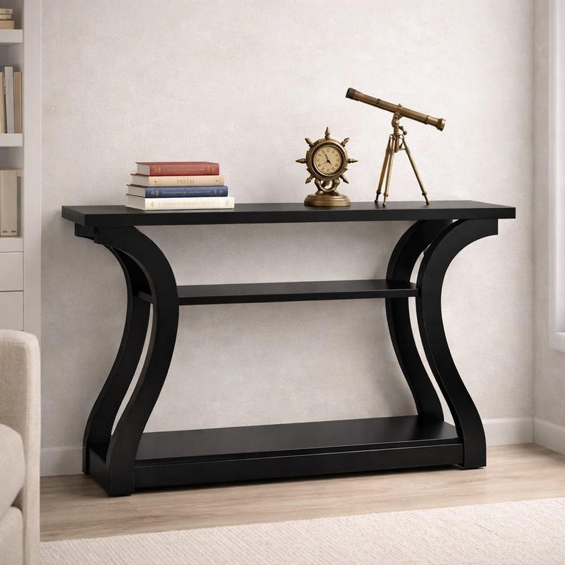 May include: A black console table with a rectangular top, a middle shelf, and a bottom shelf, featuring curved side supports. On the table sits a stack of four books and a brass-colored nautical-style clock and a brass-colored telescope on a tripod.