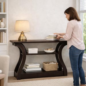 May include: A dark brown wooden console table with a curved frame, featuring a top surface, a middle shelf, and a bottom shelf. The table holds a brass-toned table lamp with a beige shade, a white box, a white bowl, and a woven basket.