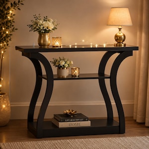 May include: A black console table with a curved frame, featuring a top surface, a middle shelf, and a bottom shelf. The table holds a gold lamp, two vases with white flowers, two candle holders, and two books titled Live Beautifully and Home.