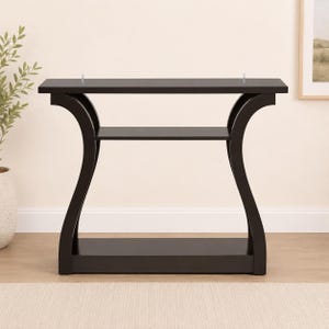 May include: A dark brown console table featuring a rectangular top, a middle shelf, and a solid base. The table is supported by two curved side panels that connect the top, middle, and bottom sections, creating a distinct open frame structure.