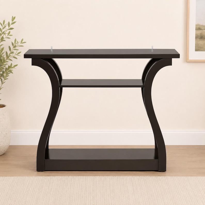 May include: A dark brown console table featuring a rectangular top, a middle shelf, and a solid base. The table is supported by two curved side panels that connect the top, middle, and bottom sections, creating a distinct open frame structure.