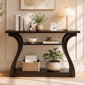 May include: A dark brown wooden console table with curved side supports and a middle shelf. The top surface holds books, a small plant, a picture frame, and a lamp. The middle shelf features a bowl and a book. The bottom shelf holds a woven basket.