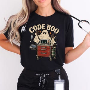 May include: A black short-sleeve t-shirt featuring a graphic print of a ghost standing behind a red medical crash cart with a heart monitor, IV pole, and oxygen tank. The text CODE BOO is printed in a beige font above the ghost illustration.