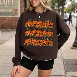 May include: A dark brown long-sleeve crewneck sweatshirt featuring a graphic print of three horizontal rows of orange jack-o'-lantern pumpkins with green leaves, small yellow butterflies, and candy corn accents on the front center.