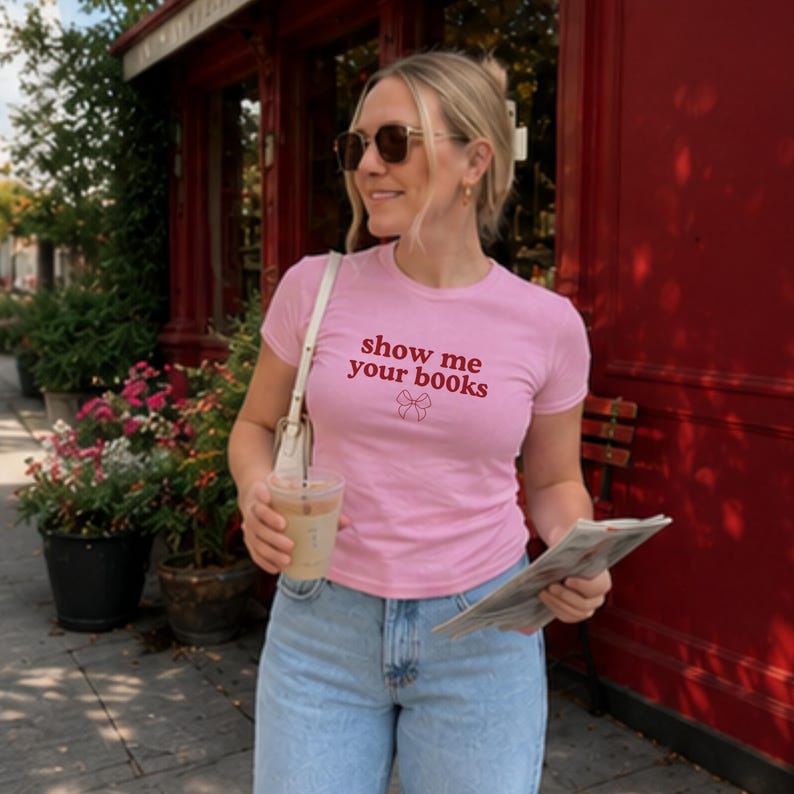 Pu&ograve; includere: Una t-shirt a maniche corte color rosa chiaro con la scritta rossa "show me your books" stampata sul petto, accompagnata da un piccolo disegno al tratto rosso di un fiocco posizionato subito sotto il testo. La maglia ha un girocollo.