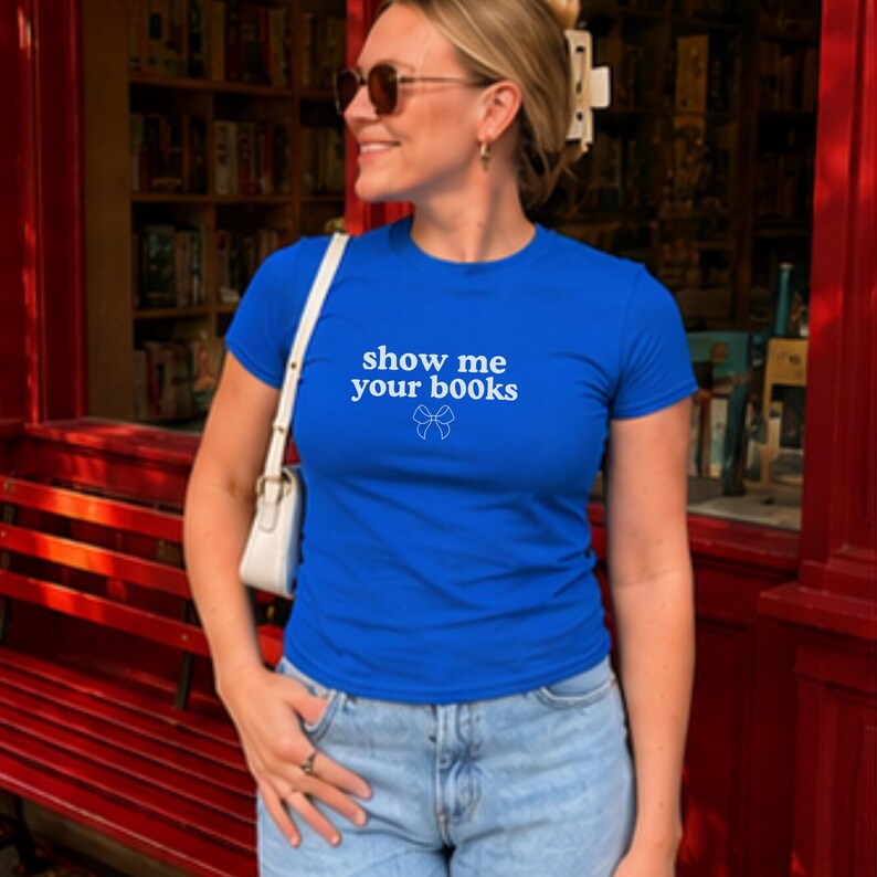 Pu&ograve; includere: Una t-shirt a maniche corte di colore blu brillante con la scritta bianca "show me your b00ks" centrata sul petto, accompagnata da un piccolo disegno a linee bianche di un fiocco posizionato appena sotto il testo.