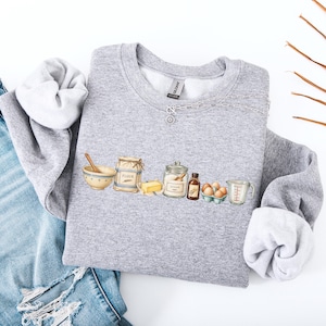 May include: A heather gray crewneck sweatshirt featuring a graphic print of baking ingredients including a mixing bowl with a wooden spoon, a bag labeled flour, a stick of butter, a sugar jar, a small bottle, brown eggs, and a measuring cup.