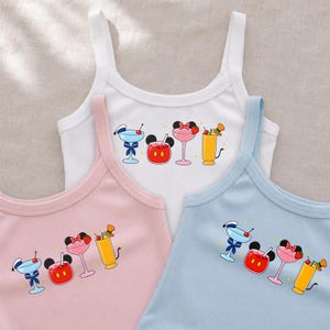 May include: Three ribbed tank tops in white, light pink, and light blue. Each top features a graphic print of four colorful cocktails decorated with iconic mouse ears, cherries, and small accessories like a blue bow, a red polka dot bow, and a hat.