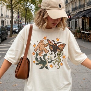 May include: A cream-colored t-shirt featuring a graphic print of a sasquatch, a moth-like creature, a sea monster, and an alien holding hands in a circle, surrounded by orange star shapes. A brown shoulder bag is worn over the side of the shirt.