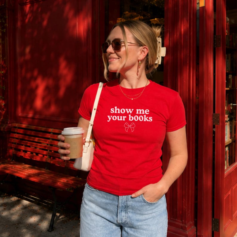 Pu&ograve; includere: Una t-shirt rossa a maniche corte con la scritta bianca "show me your books" stampata sul petto, accompagnata da un piccolo disegno a linee bianche di un fiocco situato proprio sotto il testo, abbinata a un paio di jeans azzurri.