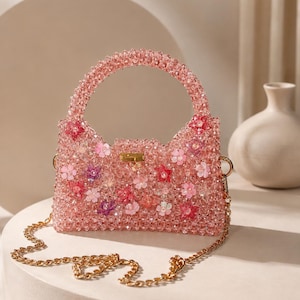 Può includere: Una borsa a mano in perline rosa con un manico superiore strutturato e una tracolla a catena color oro rimovibile. L'esterno è ricoperto di perline rosa traslucide e decorato con vari inserti a forma di fiore in tonalità rosa e rosso.