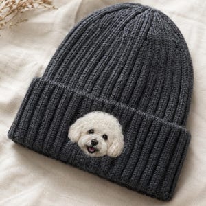 May include: A dark charcoal grey ribbed knit beanie featuring a centred, textured needle-felted patch of a white Bichon Frise dog face. The dog has black eyes, a black nose, and a small open mouth showing a pink tongue, set against the dark fabric.
