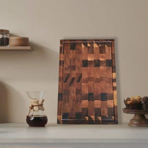 Pode incluir: Uma tábua de corte retangular de madeira com um padrão de mosaico de blocos de madeira castanha escura e clara. A tábua tem uma borda rebaixada e é exibida em uma bancada de cor clara. Uma cafeteira de vidro e um suporte de bolo de madeira também são visíveis.
